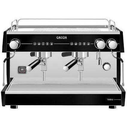 "Close-up image of a sleek Gaggia Vetro 2 Espresso machine, featuring a polished stainless steel deck with black panels on the exterior, a group head for coffee extraction, and a touch control panel of hardened glass with buttons for various brewing options."